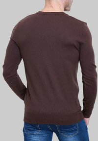 Pull marron ajusté à manches longues, en tissu texturé, avec un design à col rond, associé à un jean bleu. Vue de dos affichée.