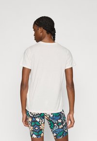 Άσπρο κοντομάνικο μπλουζάκι απεικονισμένο από πίσω, σε συνδυασμό με σορτς με σχέδια που περιλαμβάνουν μπλε, πράσινα και μπεζ λουλούδια.