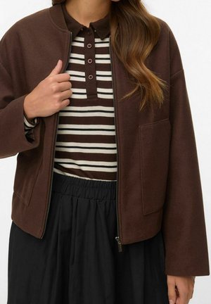 Femme portant une veste marron zippée par-dessus une chemise rayée marron et blanche à boutons et une jupe noire avec taille élastique, tenant la veste près de la fermeture éclair.