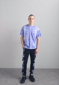 Jauns vīrietis stāv pret vienkāršu sienu, ģērbies rakstainā lilac īsām piedurknēm krekla, melnās biksēs ar lilac dizainu un melnās sporta kurpēs.