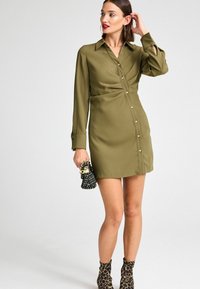 Olivengrünes, langärmeliges Hemdkleid mit Knöpfen an der Front und einer taillierten Taille. Kombiniert mit Ankle Boots im Leopardenmuster und einer schwarzen Handtasche.
