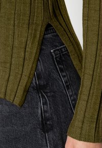 Oliwkowy sweter z ribbed, z bocznymi rozcięciami, zestawiony z ciemnoniebieskimi dżinsami, które mają kieszeń i teksturowany detal.