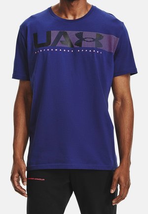 Homme portant un t-shirt bleu Under Armour avec un logo et un texte noirs et violets "Performance Apparel" sur la poitrine.
