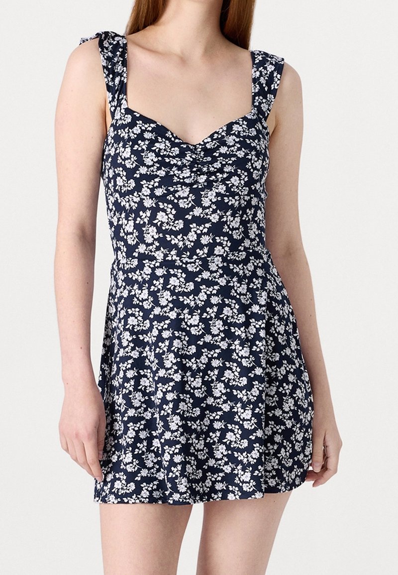 Navy blue floral dress with white patterns, featuring a fitted bodice, scoop neckline, and ruffled straps. Soft fabric and flared skirt.