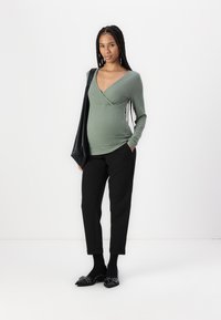 Haut long à manches ribbées vert avec un col en V, associé à un pantalon noir slim et des ballerines noires à talons pointus ornées de détails. Grand sac noir.