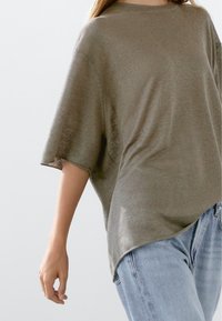 Oliwkowy, oversize sweter z miękkiej dzianiny, krótki rękaw, okrągły dekolt i zaokrąglony dół, połączony z jasnoniebieskimi dżinsami.