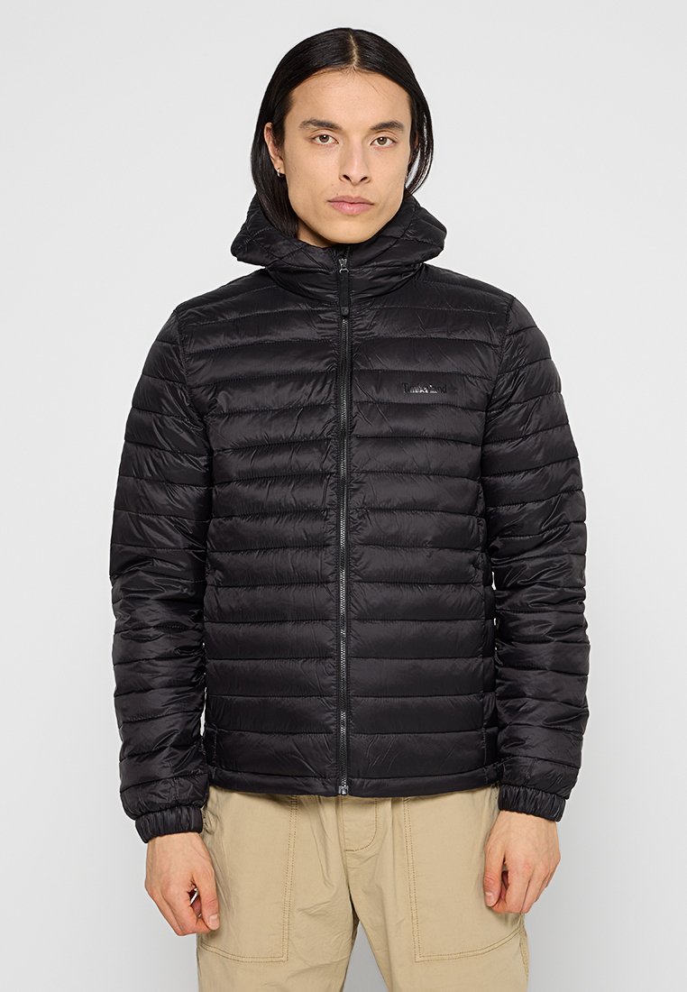 Mężczyzna z długimi ciemnymi włosami, ubrany w czarną pikowaną kurtkę z kapturem i beżowe spodnie, stojący przodem na jednolitym szarym tle.