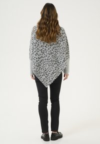 Grauer Poncho mit einem schwarzen Leopardenmuster, in dreieckiger Form und aus weichem, texturiertem Material. Getragen über schwarzen Hosen und Schuhen.
