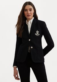 Blazer blu navy con bottoni argentati, due tasche frontali e un logo ricamato sul petto. Abbinate a una camicia bianca e pantaloni neri.
