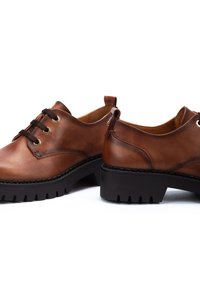 Chaussures en cuir marron avec un bout arrondi et une semelle en caoutchouc noir épais. Présentent des œillets en métal et des lacets contrastants. Texture lisse et design minimal.