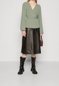 Blouse verte avec des manches bouffantes, associée à une jupe midi en cuir noir. Le modèle porte des bottes à talons noires et tient un petit sac bordeaux.