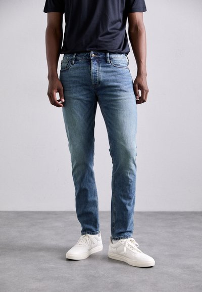 Helleblaue Jeans mit geradem Schnitt, ausgestattet mit fünf Taschen und einem dezenten Verblassen, kombiniert mit weißen Sneakers und einem schwarzen T-Shirt.