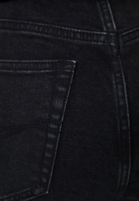 Černé džíny z denimu mají zadní kapsu, pevné švy a hladkou texturu, přičemž se zaměřují na jemný lesk tkaniny.