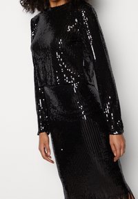 Černé šaty se třpytkami, dlouhými rukávy, kulatým výstřihem a přiléhavým střihem. Obsahují texturovanou tkaninu a hladkou sukni s třásněmi.