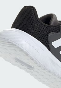 Černé síťované tenisky s strukturovaným povrchem, bílé gumové podrážky a s bočním prvkem se značkou. Mají polstrovanou patu a elegantní design.