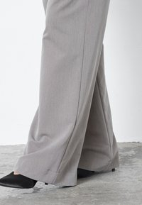 Világosszürke, bőszárú nadrág texturált anyagból, alacsony szegéllyel, fekete hegyes orrú cipővel viselve.