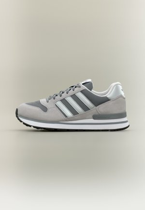 Szary sneaker wykonany z siateczki i zamszu, z trzema białymi paskami, amortyzowaną podeszwą, tonowanymi akcentami oraz wyściełanym kostką.