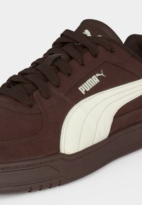 Καφέ αθλητικό παπούτσι Puma με λευκή πλαϊνή ρίγα, σχέδιο με κορδόνια και λογότυπο Puma στο πλάι και στη γλώσσα, παρουσιάζεται από λοξή κοντινή λήψη.