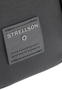 Tessuto nero texturizzato con un emblema rettangolare che presenta "STRELLSON" in lettere in rilievo e coordinate. I dettagli cuciti aggiungono profondità.