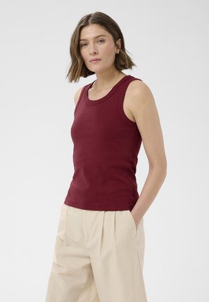 Femme aux cheveux bruns portant un top bordeaux sans manches et un pantalon beige plissé taille haute, debout avec une main dans la poche.