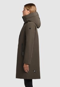 Khaaki parka bunda s vysokým golierom a veľkou kapucňou. Obsahuje zipsový bočný vrecko a akcenty loga. Hladká, vodeodolná tkanina.