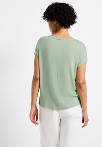 Lichtgroene top met korte mouwen, comfortabele pasvorm, zachte textuur en ronde halslijn. De achterkant heeft een middelste naaddetail. Gemaakt in combinatie met witte broek.