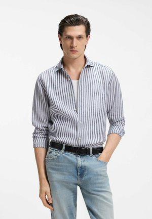 Junger Mann mit zurückgekämmtem Haar, der ein vertikal gestreiftes Hemd mit Knöpfen, ein weißes Unterhemd, hellblaue Jeans und einen schwarzen Gürtel trägt.