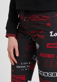 Czarne dresy z kolorowym nadrukiem o tematyce podróżniczej, zawierające nazwy miast i informacje o lotach w czerwonym i białym tekście. Teksturowany materiał z elastanem.