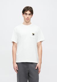 Λευκό βαμβακερό T-shirt με κοντά μανίκια, με τσέπη στο στήθος διακοσμημένη με γραφικό βουνό. Φοριέται με γκρι παντελόνι με κορδόνι στη μέση.