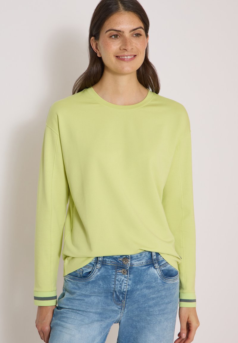 Femme aux cheveux foncés souriant, portant un haut vert clair à manches longues et un jean bleu denim sur un fond clair uni.