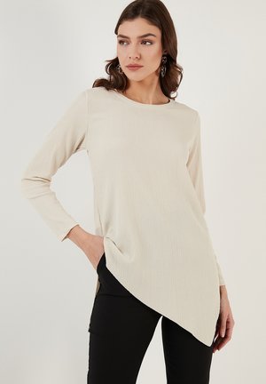 Tunic beige a maniche lunghe con superficie testurizzata, caratterizzata da un colletto rotondo e un orlo asimmetrico. Abbinamento con pantaloni neri e tasche visibili.