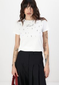 Camiseta de algodón blanca con detalles plateados adornados y un diseño en cursiva. Mangas cortas, cuello redondo, combinada con pantalones negros con pliegues.