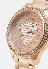 Eine roségoldene Uhr mit einem glitzernden Zifferblatt mit ausgeschnittenen Streifen, einem mit Kristallen besetzten Gehäuse und einem polierten Metallarmband.
