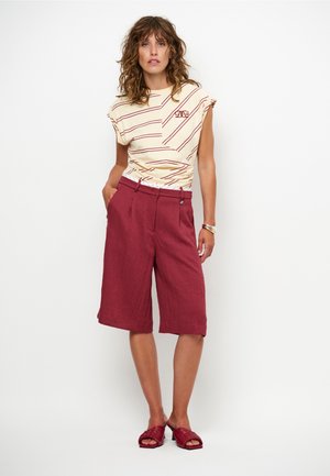 Crèmekleurig topje met rode strepen en korte mouwen, gecombineerd met bordeauxrode shorts tot op de knie en rode geweven slippers. Accent met een zilveren armband.