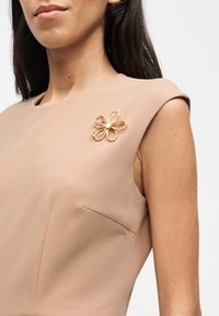 Γυναίκα με αμάνικο μπεζ φόρεμα και χρυσό καρφίτσα σε σχήμα λουλουδιού στον αριστερό ώμο, σε απλό λευκό φόντο.