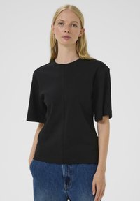Il modello indossa una maglietta nera a maniche corte, rivolto di fronte con un'espressione neutra, abbinata a pantaloni di jeans azzurri su uno sfondo bianco.