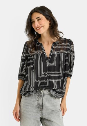 Femme aux longs cheveux bruns portant un chemisier noir avec des lignes géométriques blanches et un jean gris taille haute, souriante avec la tête légèrement tournée.