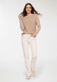 Beżowy sweter z dzianiny z okrągłym dekoltem, w połączeniu z białymi prostymi spodniami dżinsowymi i beżowymi sneakersami, stojący na jasnym tle.