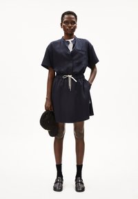 Marineblauwe korte mouwen button-up jurk met een elastische tailleband en een trekkoord. Bevat een borstzak en is gecombineerd met zwarte instapschoenen en sokken.