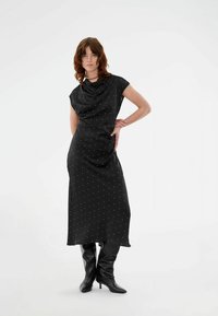 Femeie cu păr până la umeri, purtând o rochie midi neagră cu buline și cizme negre cu toc, stând cu o mână pe șold pe un fundal alb.