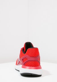 Zapatilla atlética roja con parte superior de malla texturizada, soporte para el talón sintético y suela blanca. Presenta tres franjas blancas en los lados.