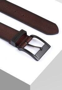 Bruine leren riem met een gladde textuur, voorzien van een matte zwarte metalen gesp. De riem loopt taps toe aan het einde met gaatjes voor een verstelbare pasvorm.