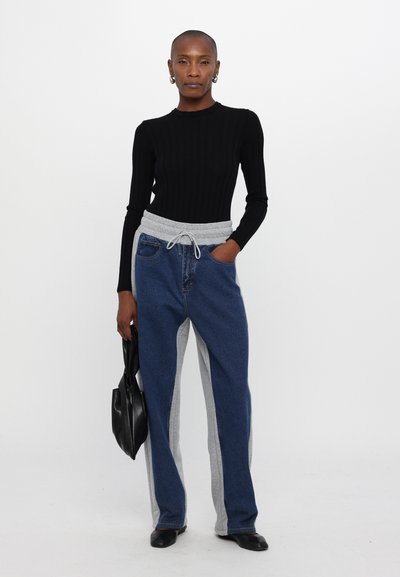 Top negro de manga larga acanalado combinado con pantalones de mezclilla azules que tienen paneles laterales grises y cordón ajustable. La modelo sostiene un bolso negro.