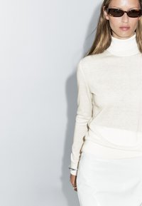 Pull col roulé crème en maille douce, associé à une jupe blanche ajustée. Modèle portant des lunettes de soleil foncées, avec des cheveux longs et raides.