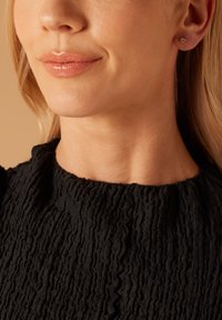 Gouden stud oorbel in een ronde vorm. Zwarte, gestructureerde stof bovenkant met een hoge, gerimpelde halslijn. Glad huidtype met een neutrale lipkleur.