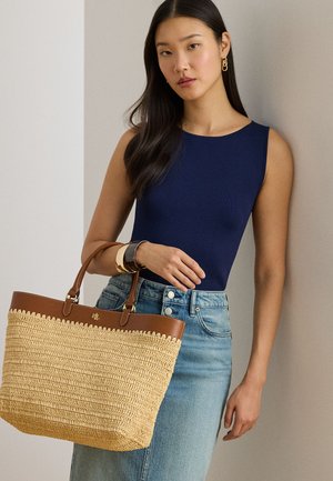 Naine, kes kannab mereväe sinist varrukateta topi ja teksaseelikut, hoides suurt kootud kandekotti, millel on pruun nahast ääris, ning käel kullast käevõrusid.