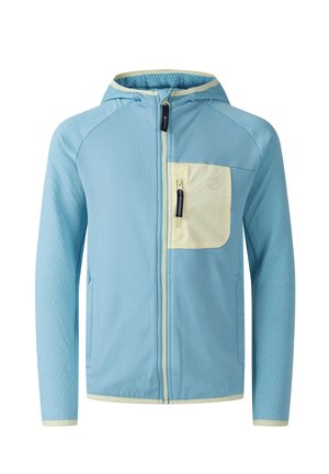 Ein junges Mädchen trägt eine blaue Jacke, dunkelblaue Shorts und Laufschuhe, hält eine kleine lila Tasche, lächelt und schaut zur Seite.