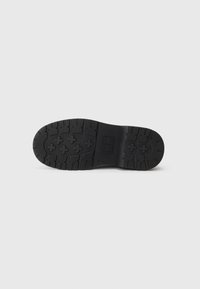 Semelle en caoutchouc noir avec un motif texturé, présentant divers motifs de crampons pour l'adhérence et la stabilité. Logo de marque rectangulaire au centre.