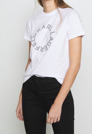 Femme portant un t-shirt blanc à manches courtes avec le texte « Karl Lagerfeld » formant un cercle, associé à un pantalon noir taille haute.