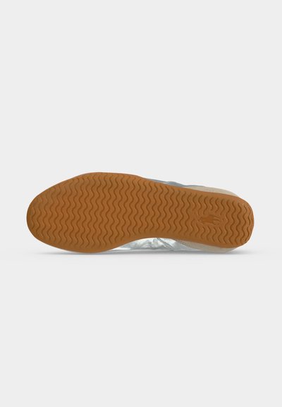 Semelle de chaussure en caoutchouc brun clair avec un motif de bande de roulement ondulé vue par dessous sur un fond gris neutre.